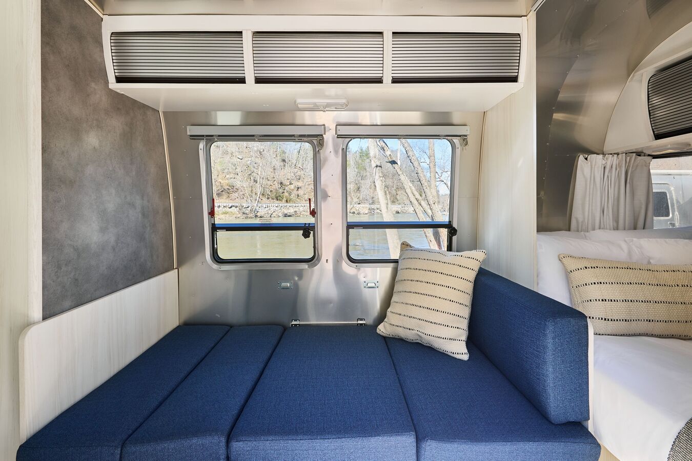 Airstream trailer seating area converted into a flat bed, positioned beside windows with outdoor views and adjacent to a rear sleeping nook.