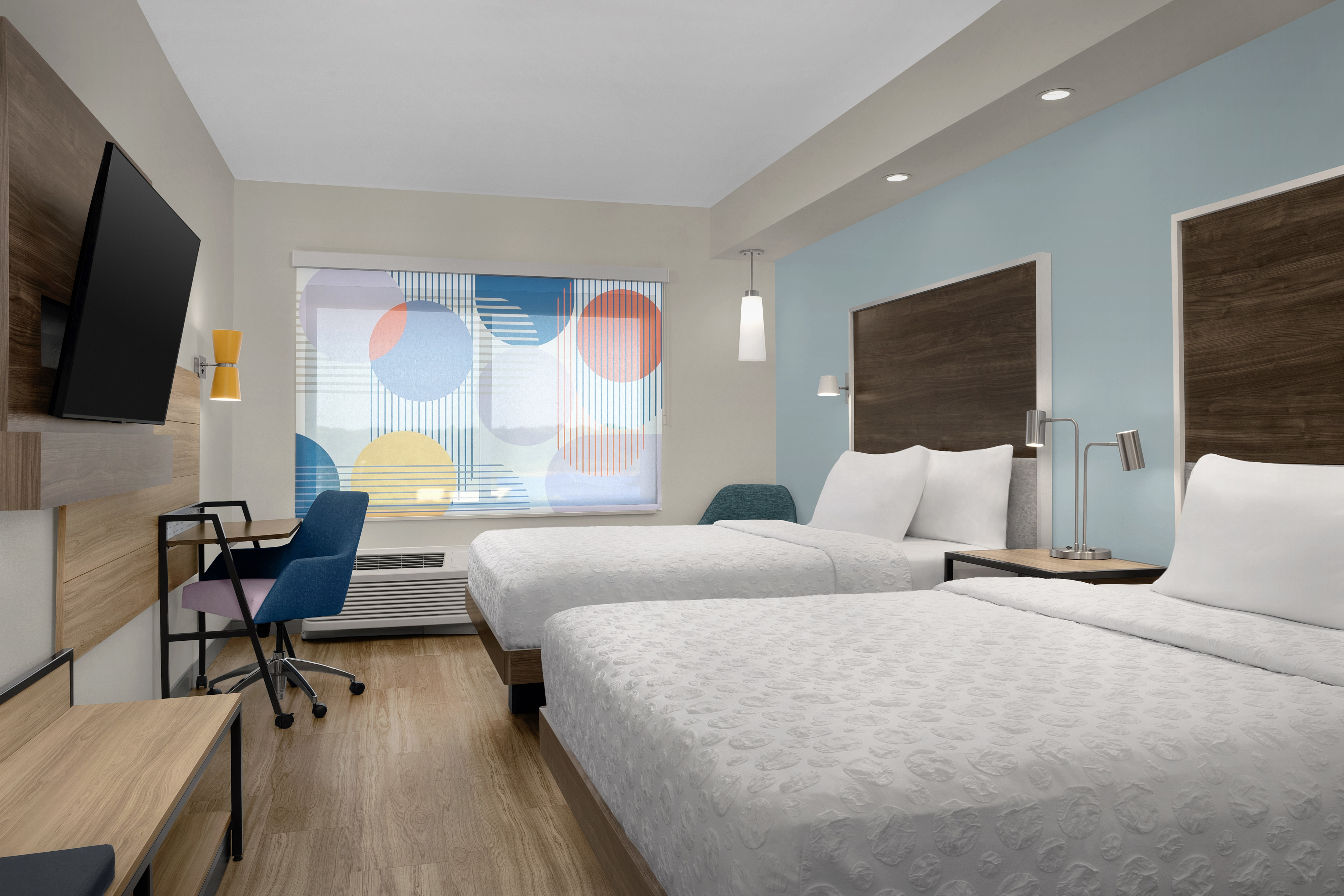 Guestroom with two queen beds, flatscreen tv, desk with chair and light filtering through windows with colorful blinds.