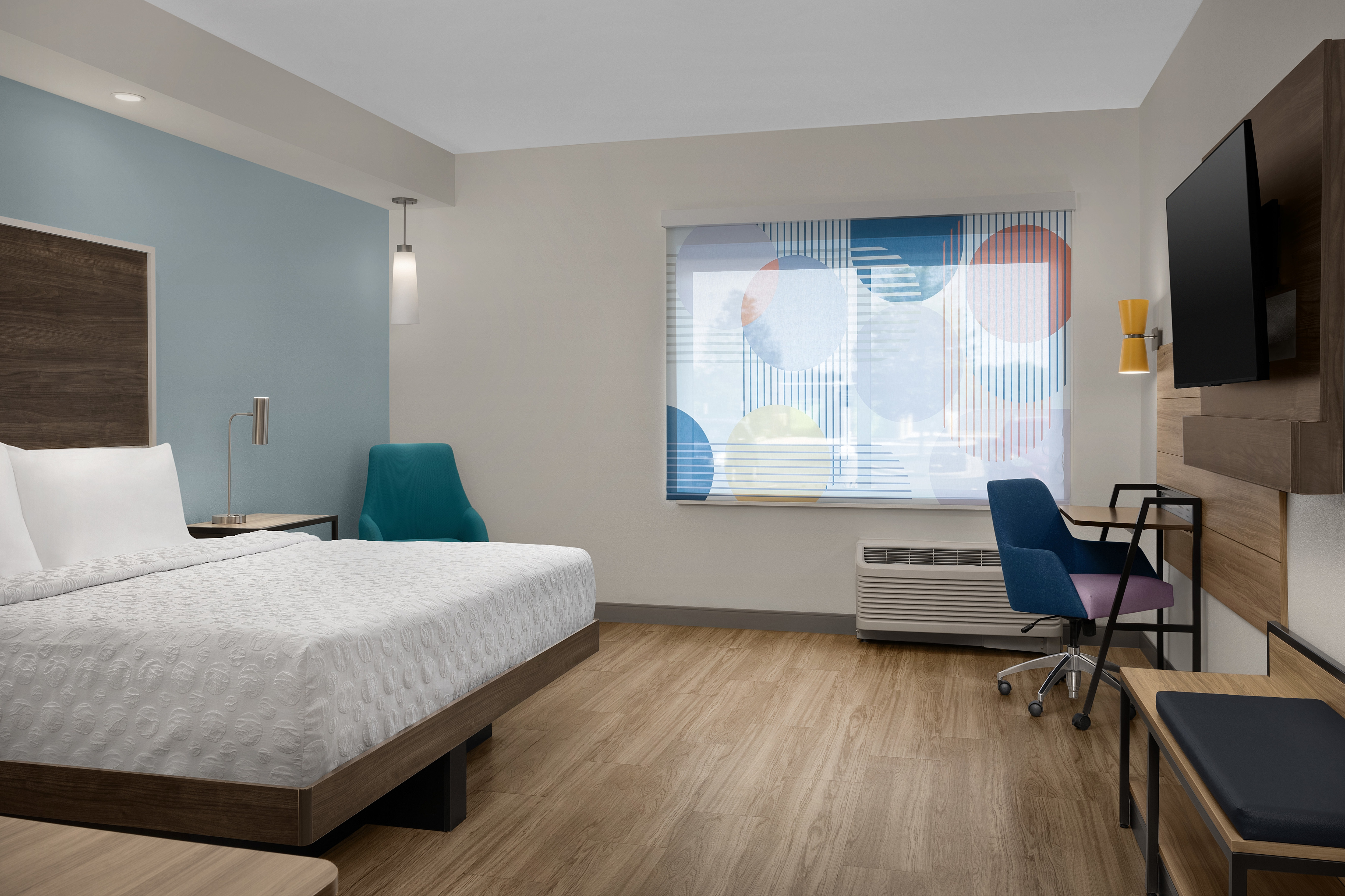 Spacious guestroom with king size bed, tv, desk, desk chair, arm chair and light filtering in through window covered with colorful blinds.