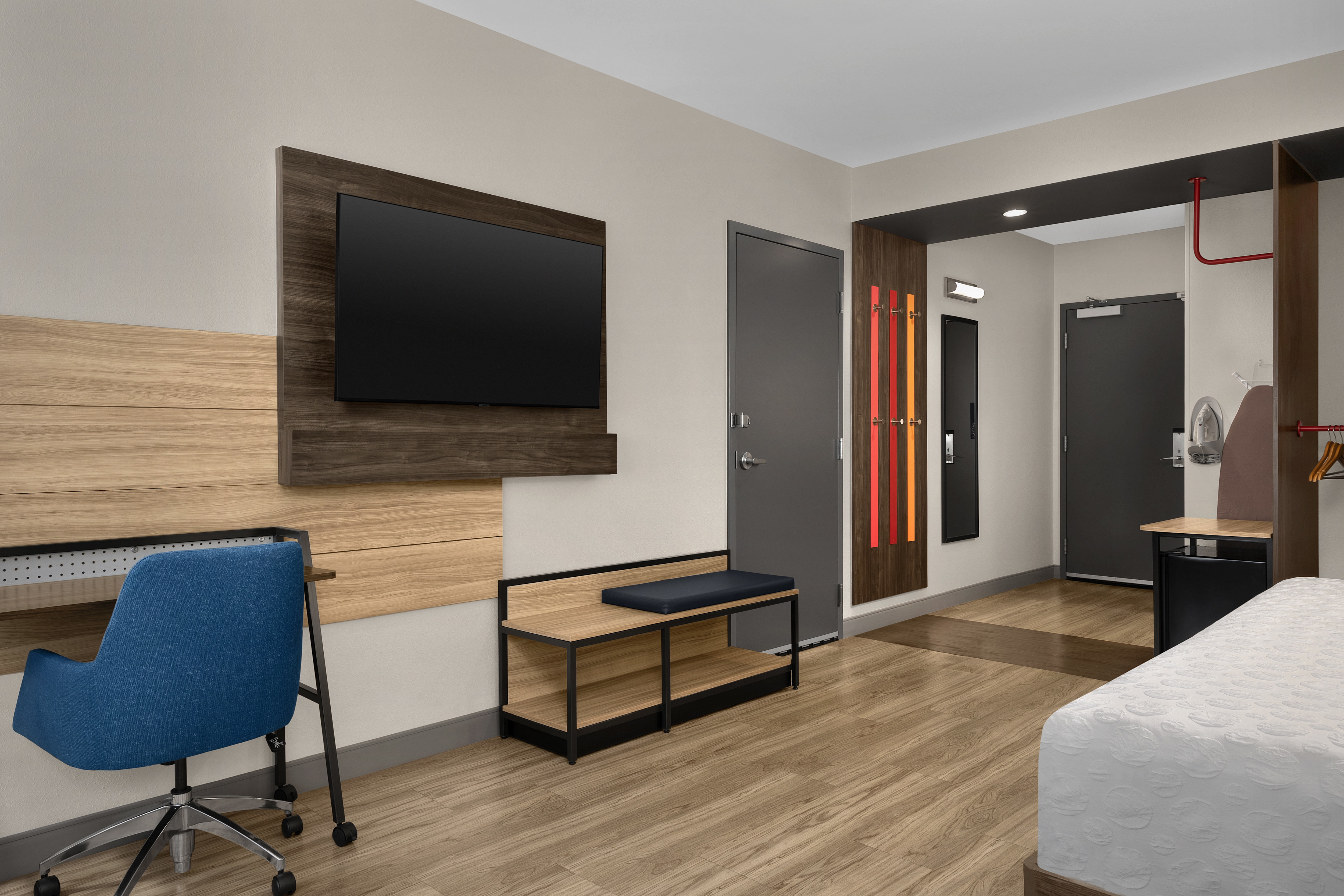 Lounge area of modern  guestroom with wooden flooring, desk, flatscreen tv and bed with view to entrance.