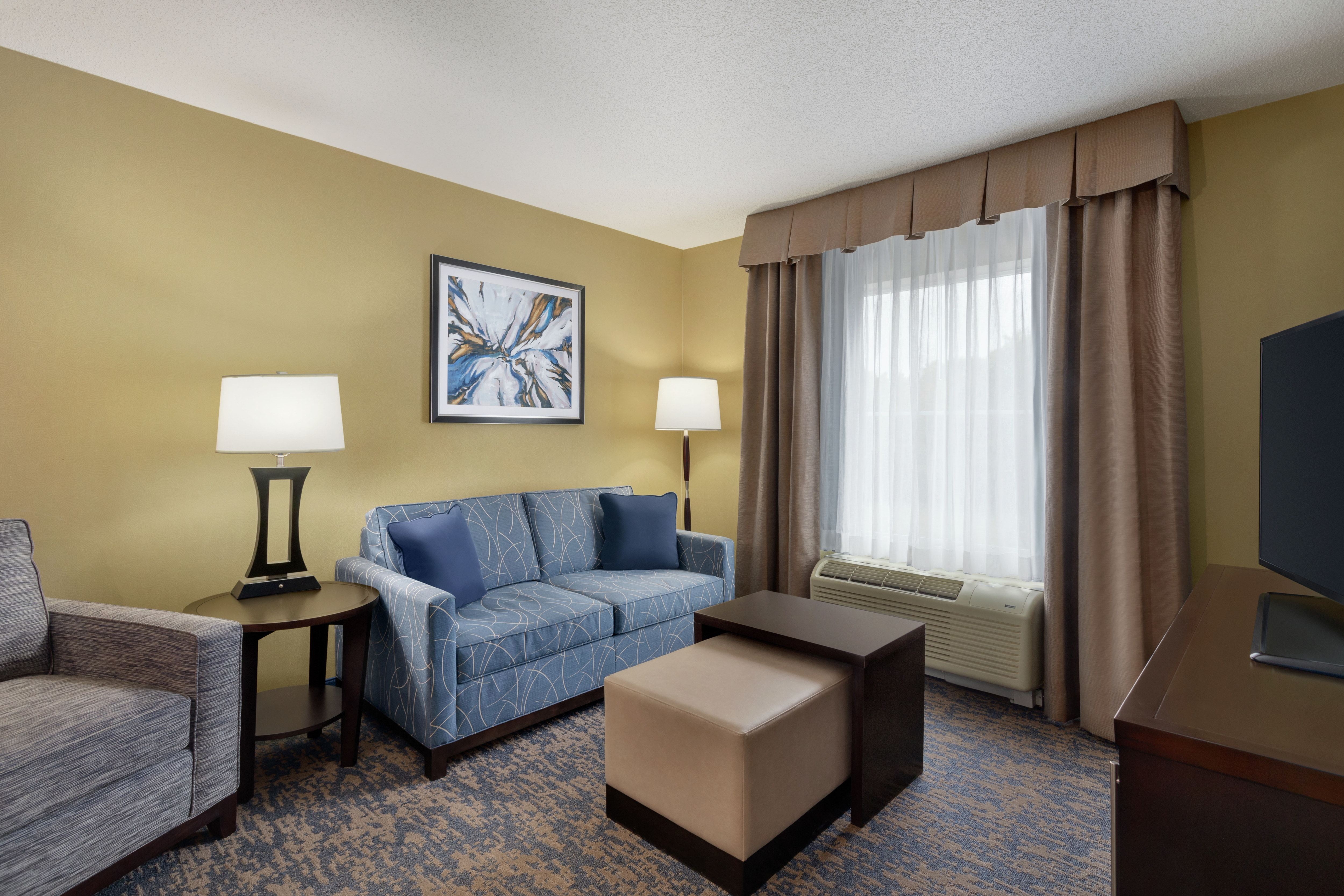 Bright lounge area in suite featuring comfortable seating, TV, and air conditioning unit by window. 