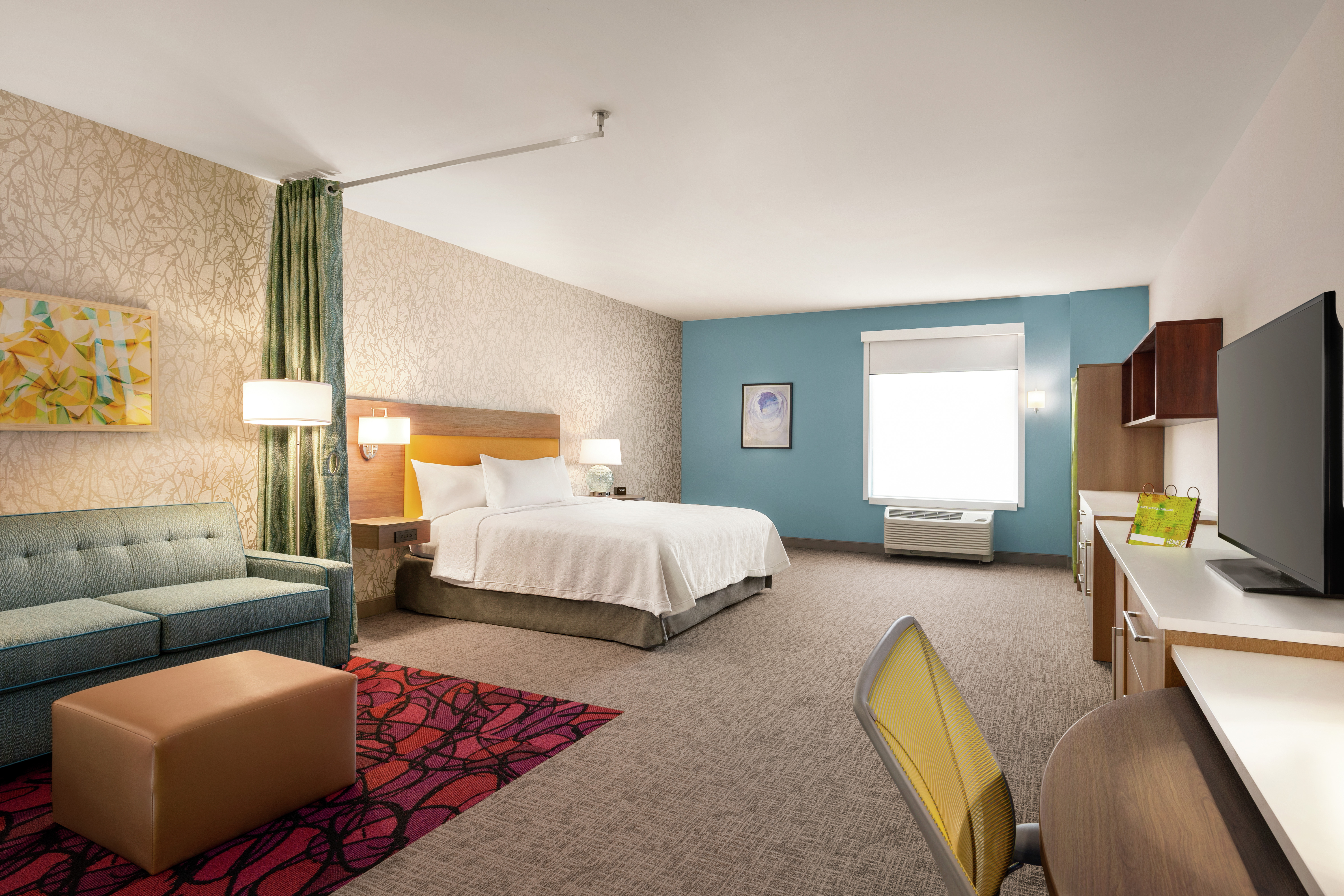 Spacious accessible studio suite featuring sofa, work desk with ergonomic chair, TV, comfortable king bed, and air conditioning unit by window.