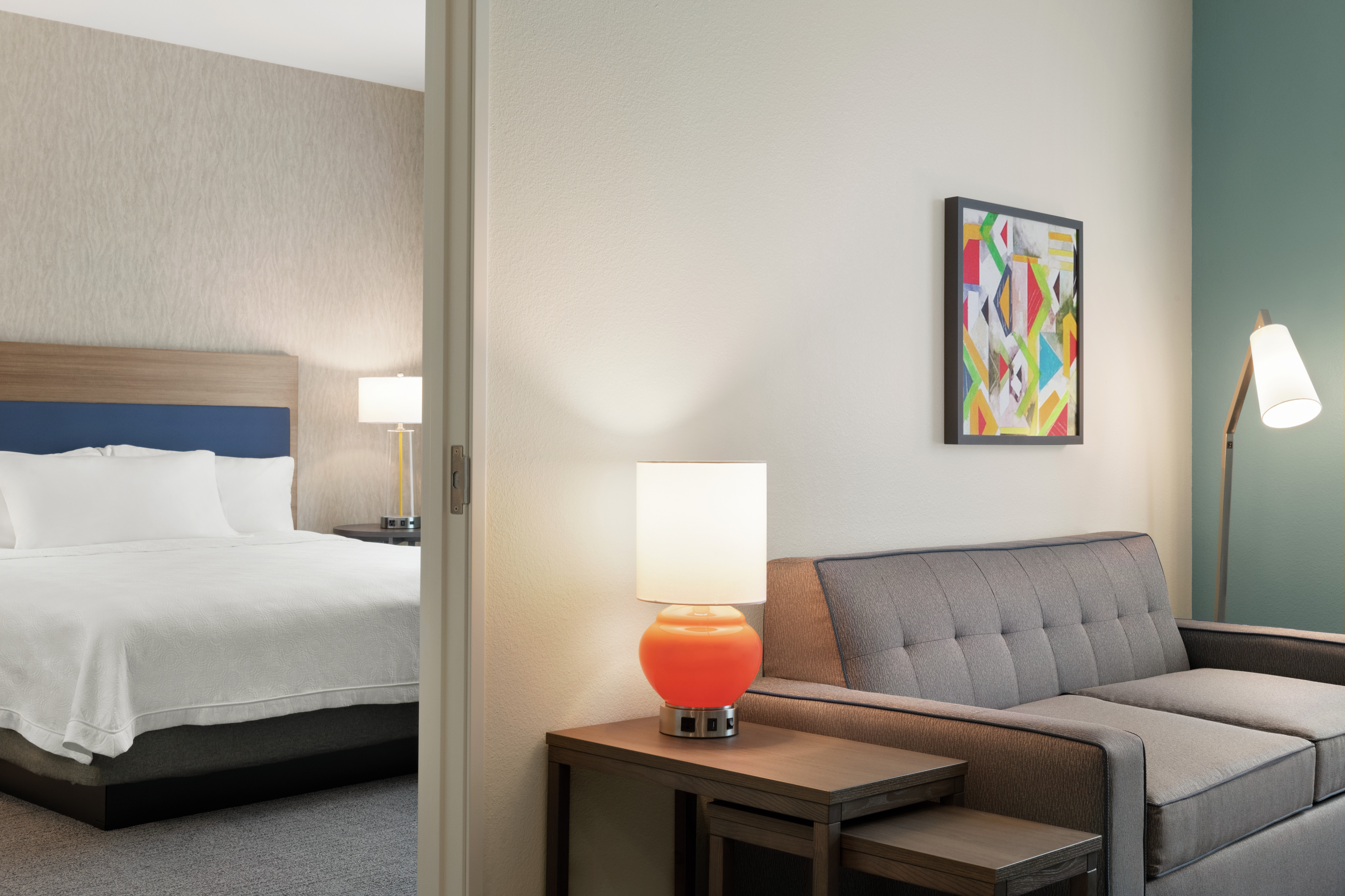Bright lounge area in suite featuring sofa, side table with lamp, geometric framed art on wall, and view into private bedroom.