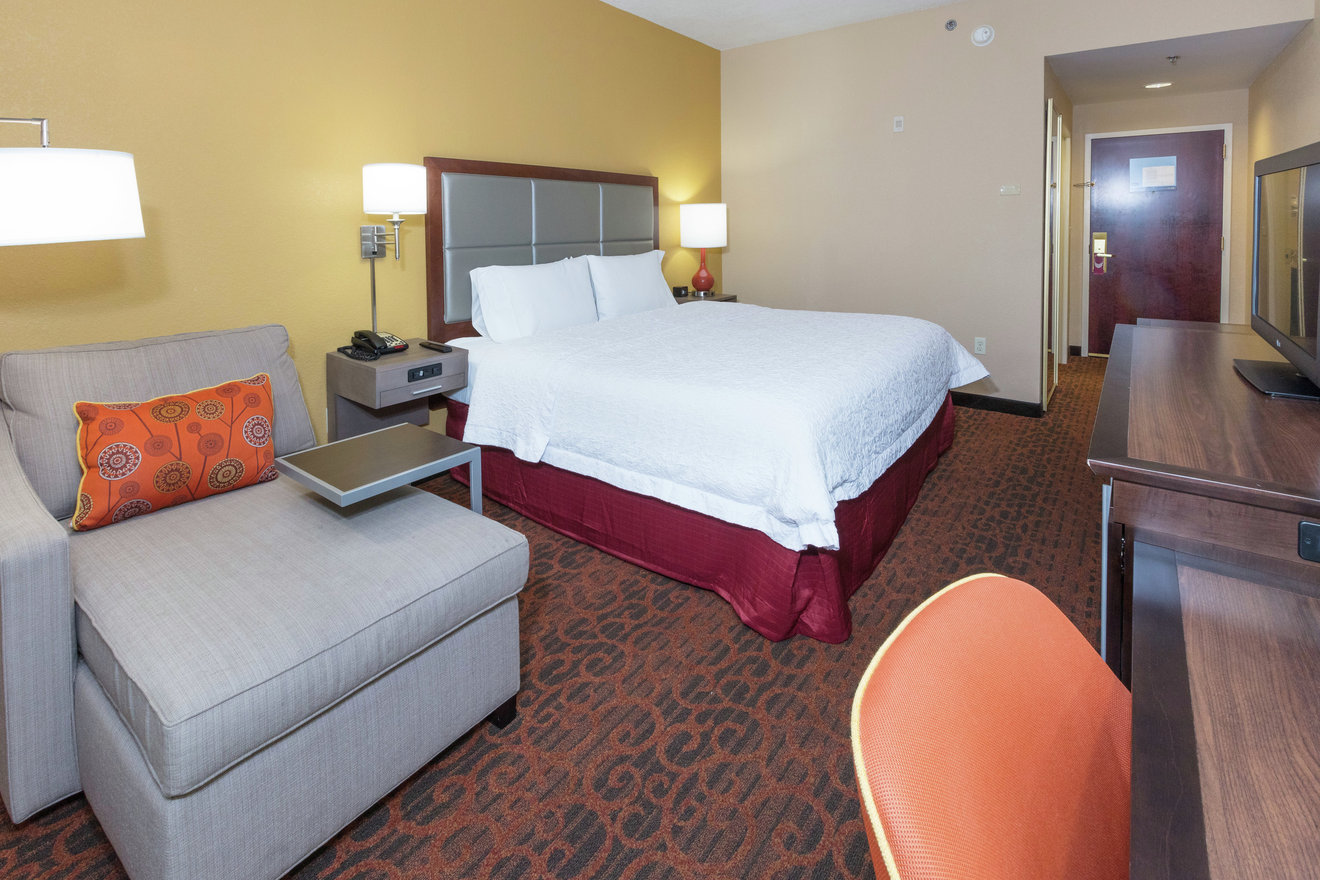Guest Room With King Bed Between Illuminated Lamps, Entry,TV, Work Desk, and Armchair
