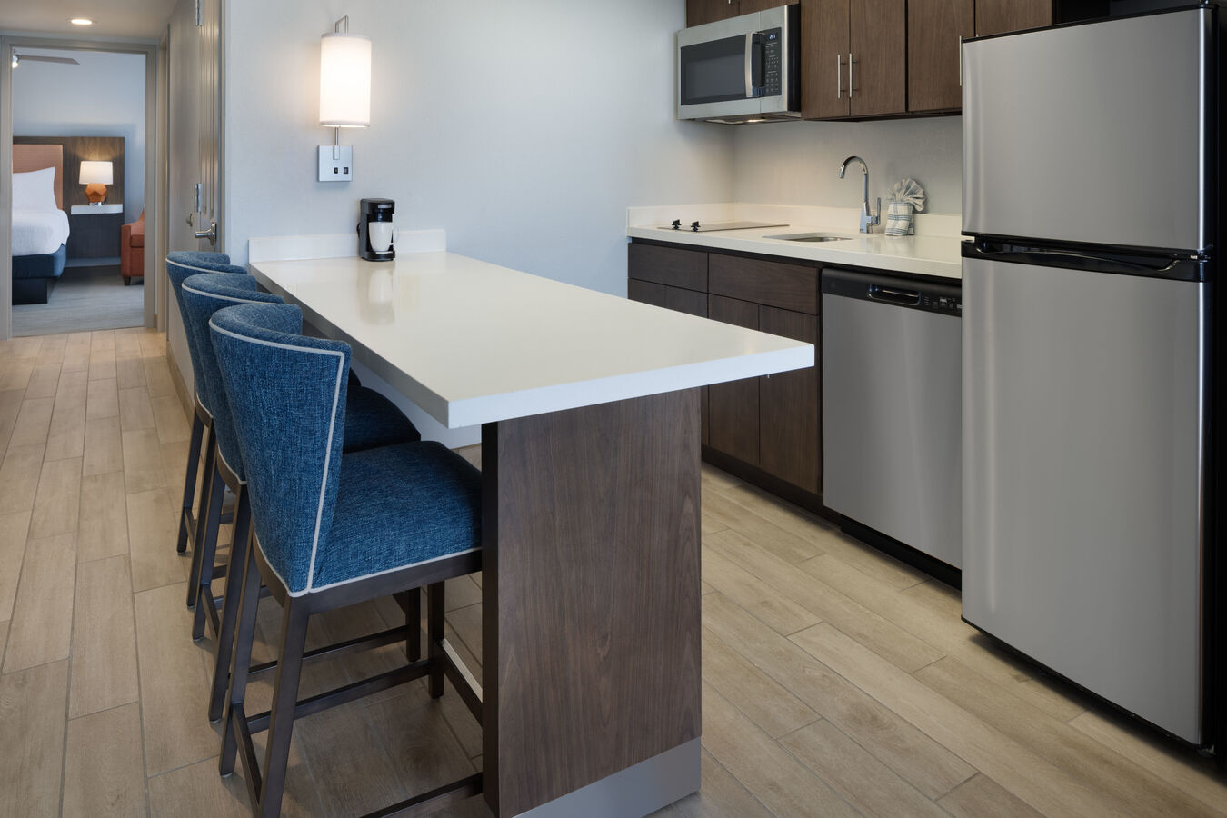 Kitchen in a hotel room with full-size appliances and a bar with four stools and down the hall an open door to bedroom.