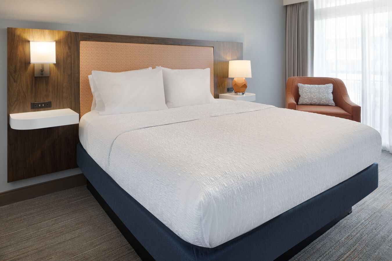 King bed with wall-mounted floating shelf on both sides of the bed, and a wall of windows in a hotel room.