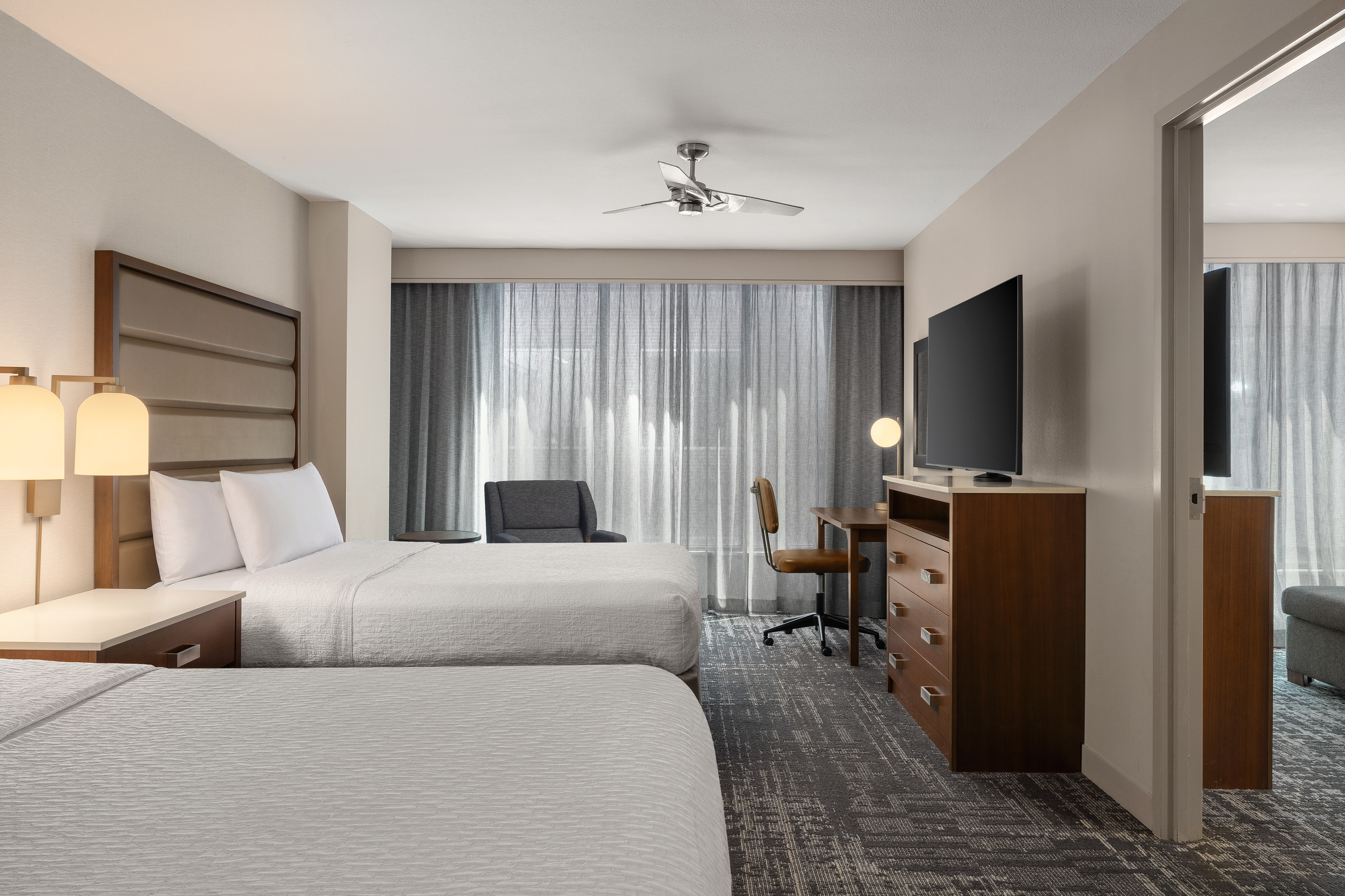 Two beds in a guest room with a nightstand, a work desk area, and a television on a dresser with views into the lounge area. 